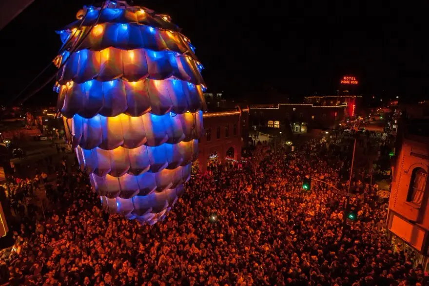 A large, illuminated pinecone-shaped hot air balloon floats over a dense crowd at night, its scales glowing blue and orange…