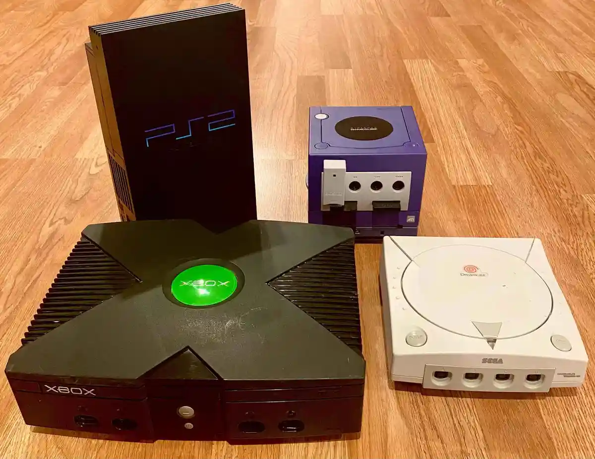 Four gaming consoles from the early 2000s are arranged on a wooden floor: a black Xbox with a glowing green logo, a white…