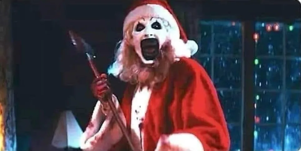 A person dressed as a Santa Claus with a white painted face, black eye makeup, and a bloody mouth is screaming while holding…
