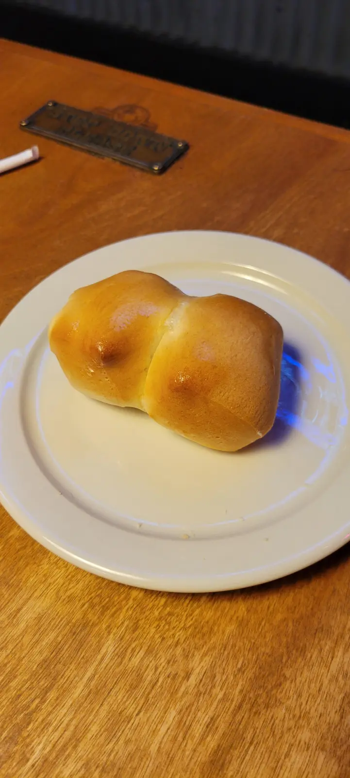 A golden-brown, glossy dinner roll sits on a white plate atop a wooden table, its shape resembling two rounded halves…