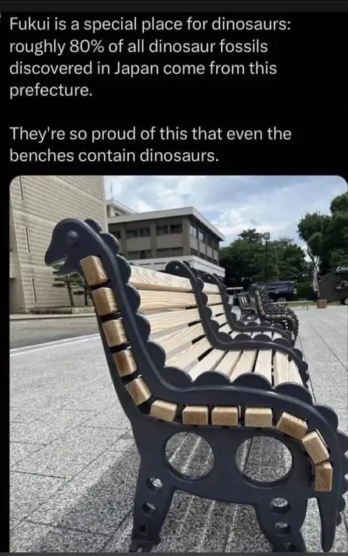 An outdoor photo shows a row of wooden benches with black metal frames shaped like dinosaurs, featuring a dinosaur head at…