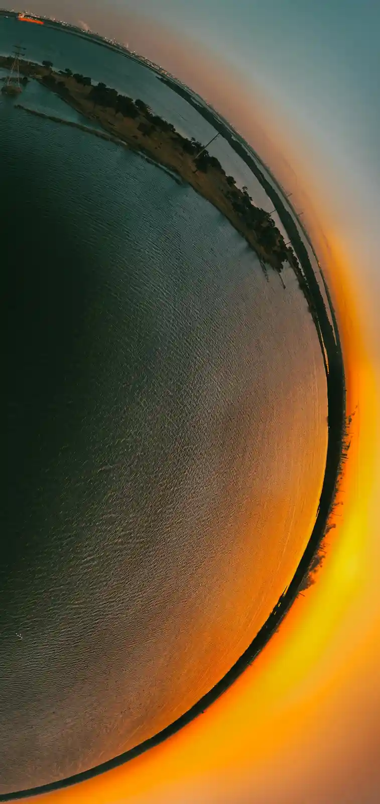 A fisheye lens photo shows a curved horizon over water at sunset, with orange and blue tones, distant land, and a ship. The…