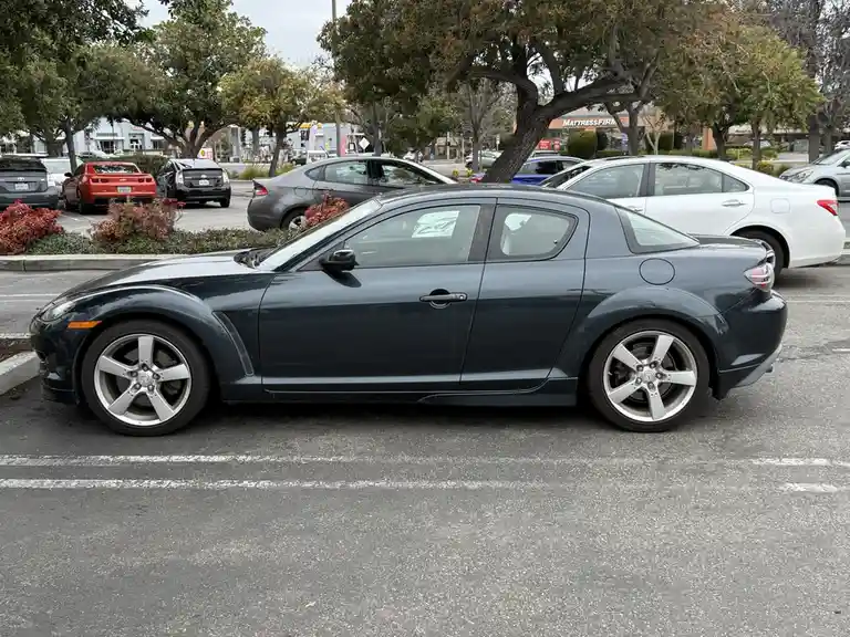 A dark gray Mazda RX-8 is parked in a public lot, viewed from the side. Other cars and trees are visible in the background…