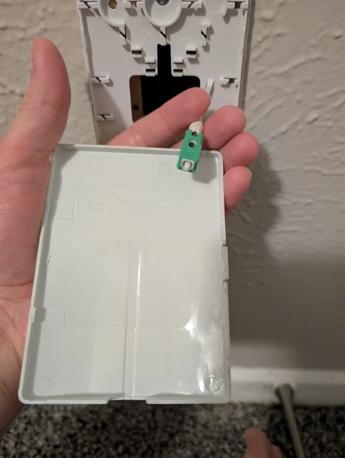 A hand holds a white plastic electrical device, likely a smart home hub or communication module, with a green wire connector…