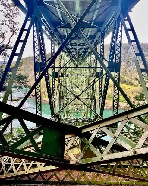 A low-angle photo looking up through the green-painted steel trusses of a large bridge, showing the intricate lattice…