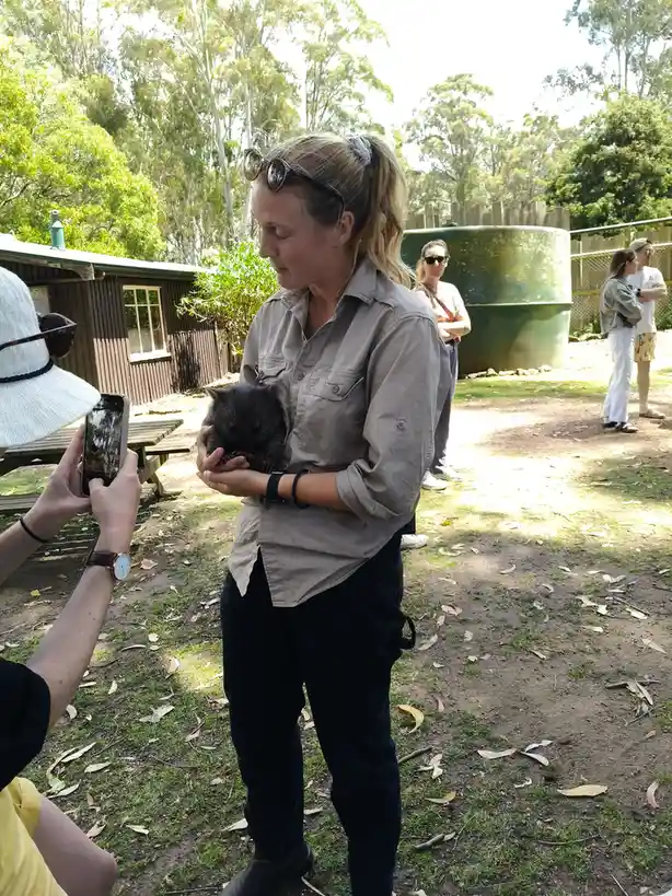 A woman in a khaki shirt holds a small, dark-furred wombat while another person takes a photo of them with a smartphone. The…