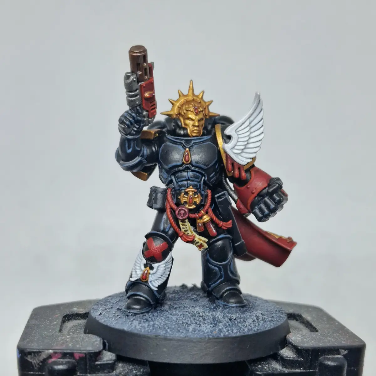A painted miniature of a Space Marine from Warhammer 40,000, wearing black armor with red and gold accents, white wings, and…