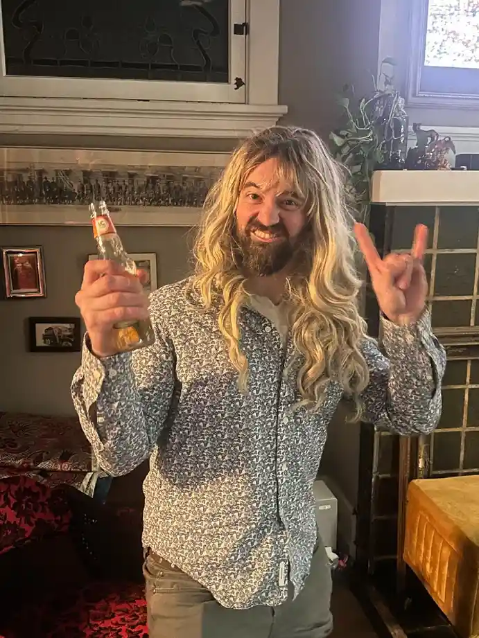 A man with long blonde hair and a beard, wearing a patterned button-up shirt, holds a beer bottle in one hand and makes a…