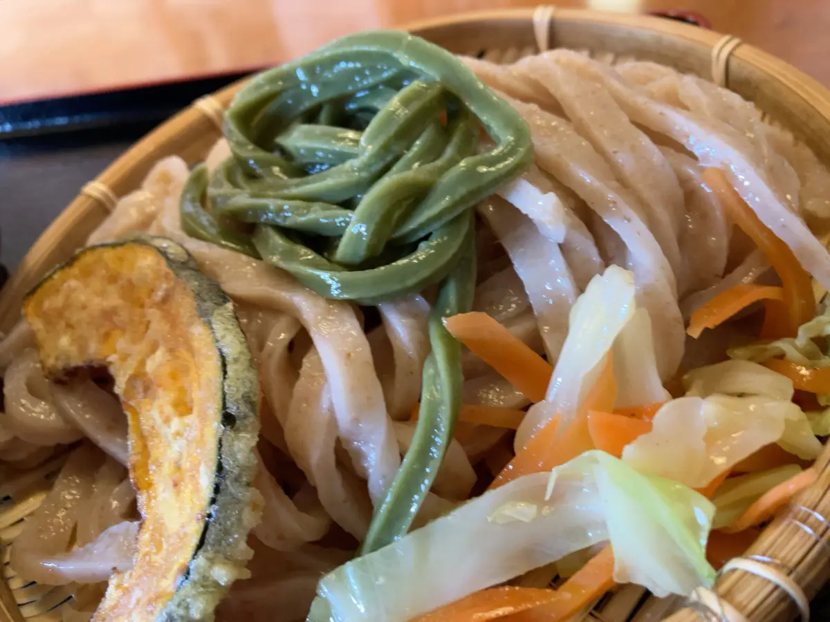 a close-up photo of a bamboo basket filled with udon noodles, topped with a swirl of green bean garnish, shredded carrots…