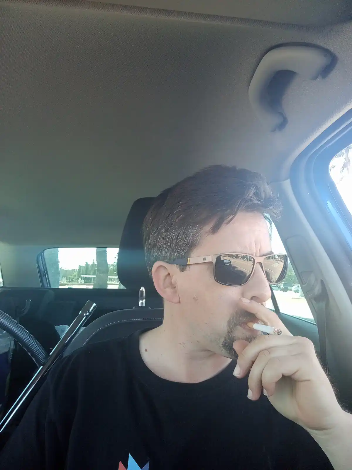 A man with dark hair and a goatee, wearing sunglasses and a black t-shirt, sits in the driver's seat of a car while holding…