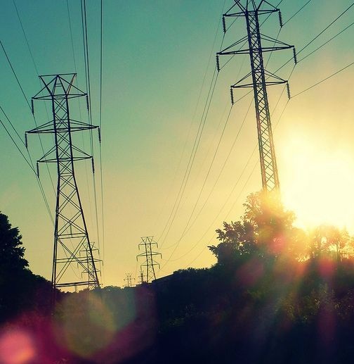 A scenic photo of tall electricity pylons silhouetted against a bright, hazy sunset with lens flare. Overlaid text reads…