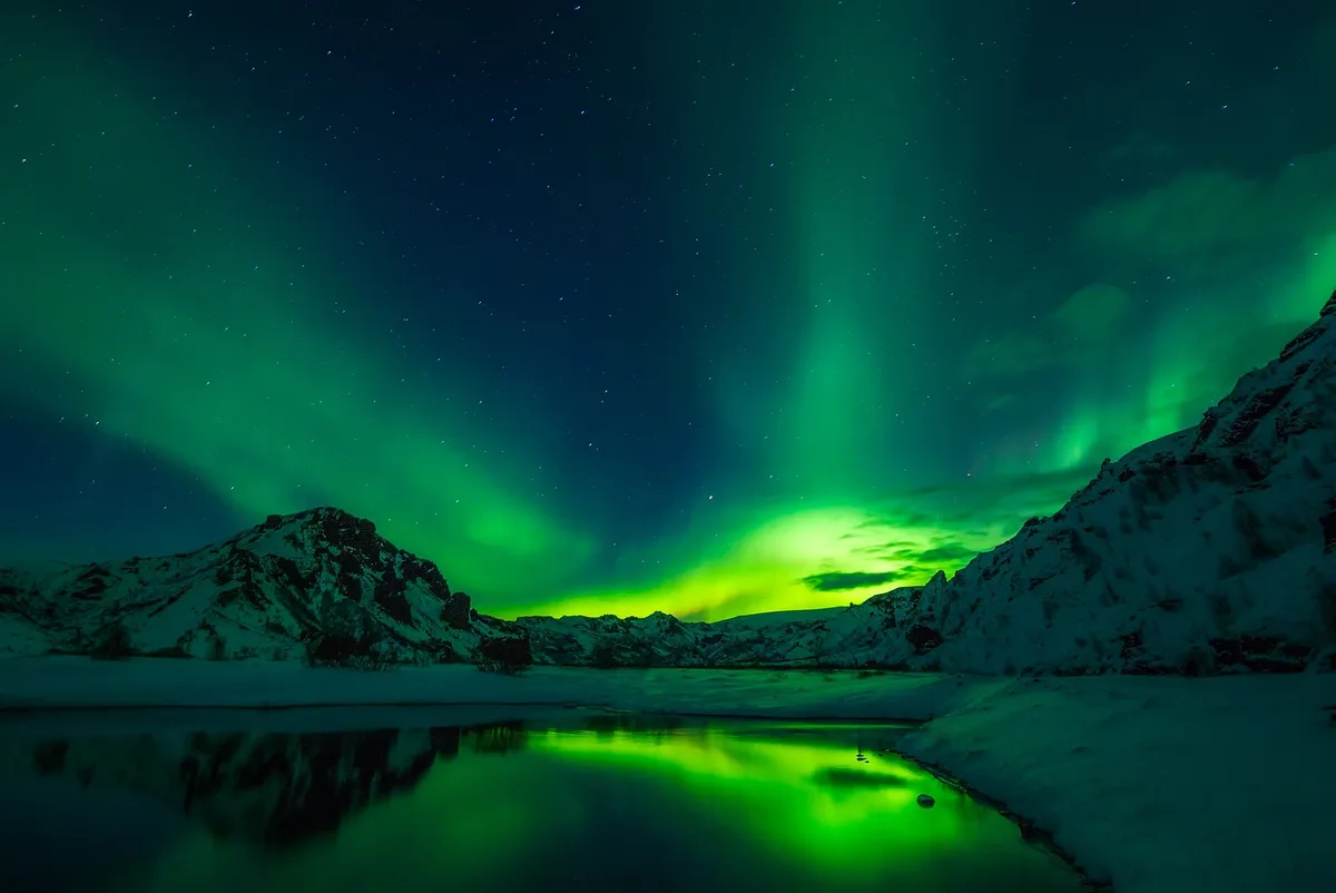 A stunning photograph of vibrant green northern lights dancing across a starry night sky over snow-covered mountains and a…