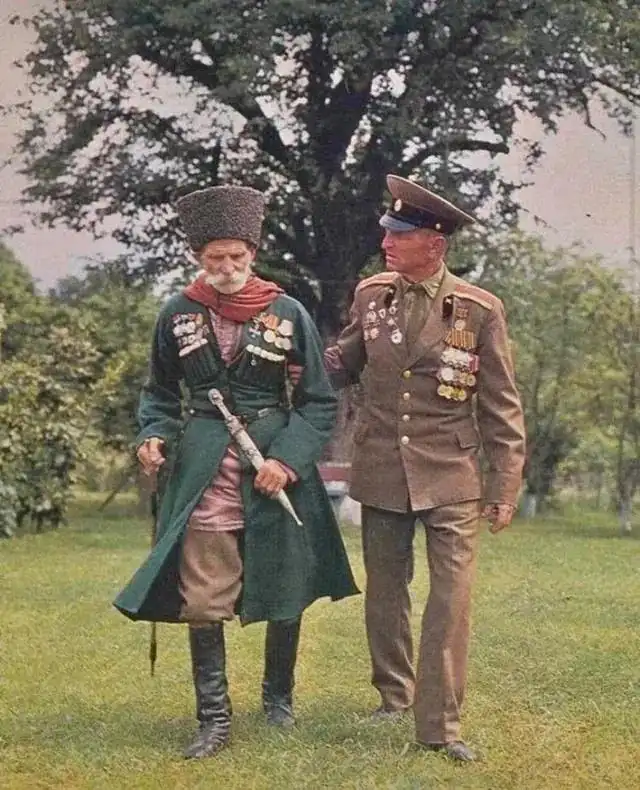 Two men in military-style attire stand outdoors on grass under a tree. The man on the left wears a traditional hat, green…