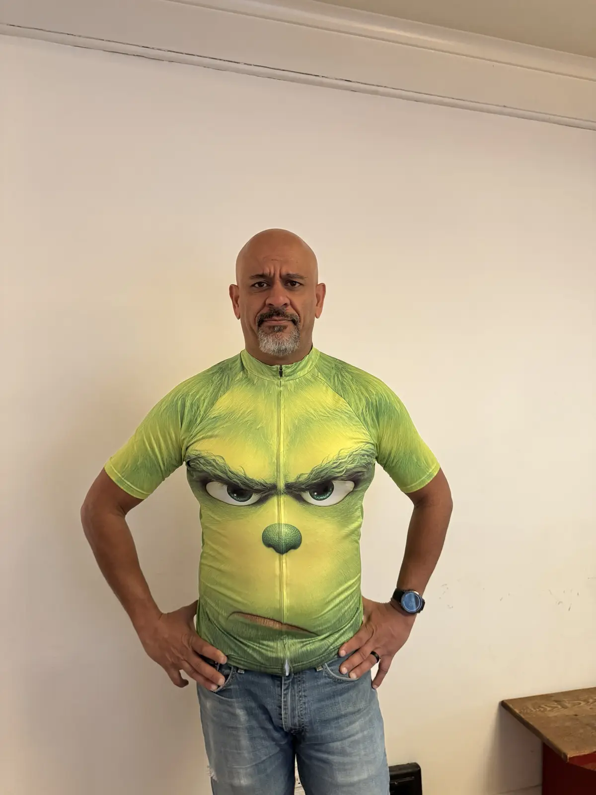 A bald man with a goatee stands with hands on hips wearing a bright green cycling jersey that features a large, angry face…