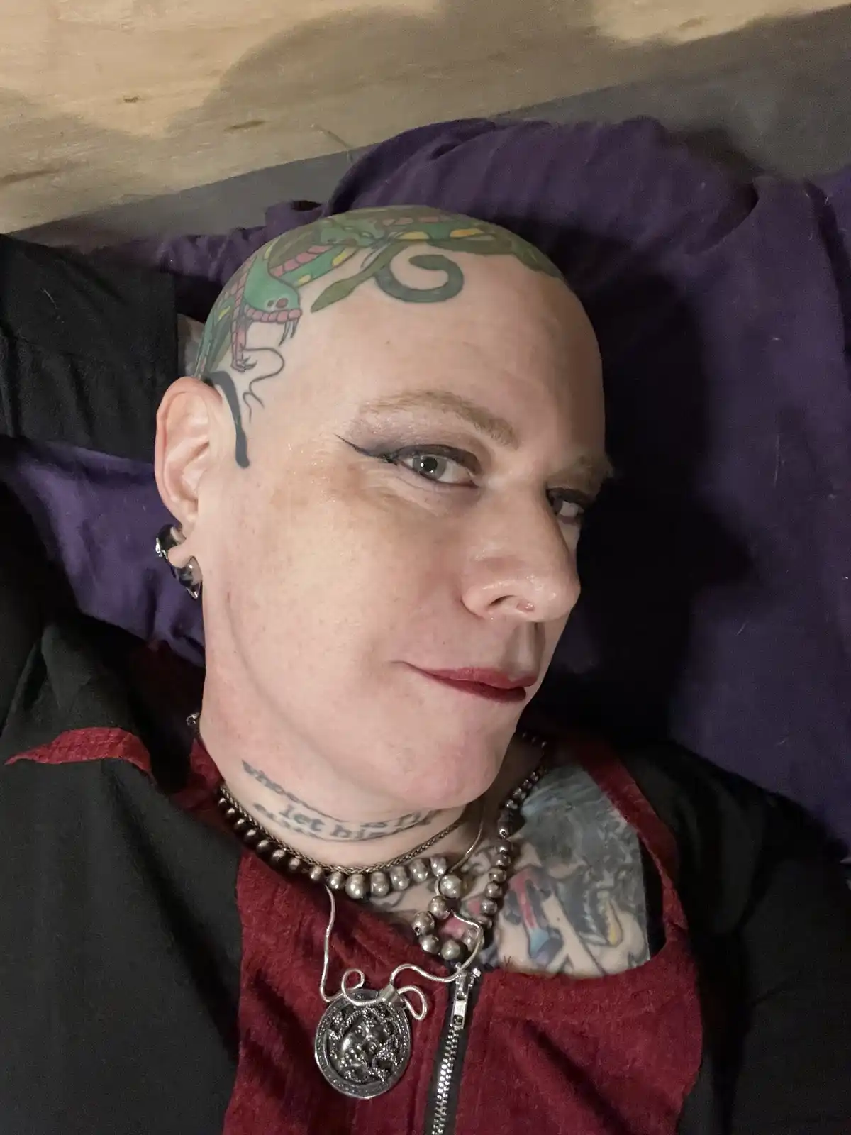 A person with a shaved head and a large green snake tattoo on their scalp lies on a purple pillow, wearing a black and red…