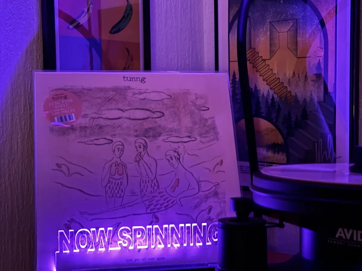 A vinyl record by artist tunng, featuring a sketch of three figures in swimwear, is displayed under purple ambient lighting…