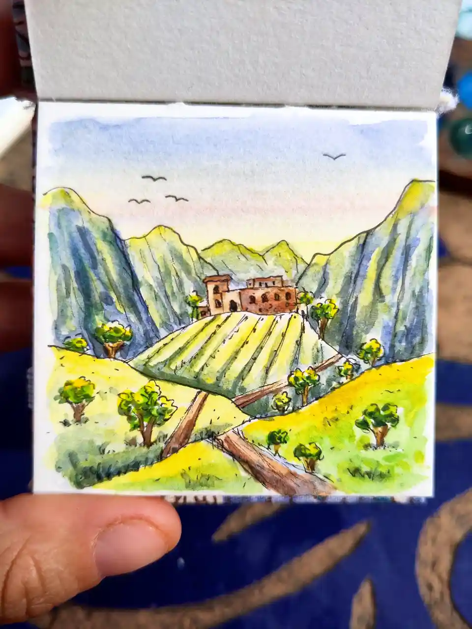 a hand holds a small watercolor painting depicting a scenic landscape with green and yellow rolling hills, a brown building…