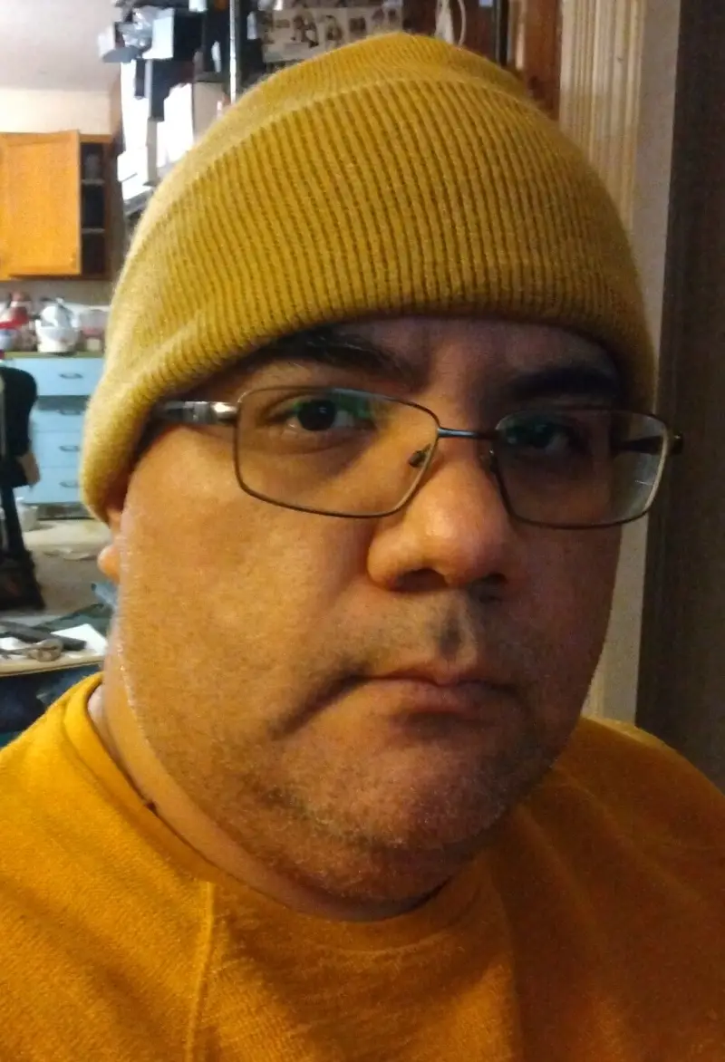 A man wearing a yellow beanie and glasses stares directly at the camera with a neutral, slightly weary expression. He is…