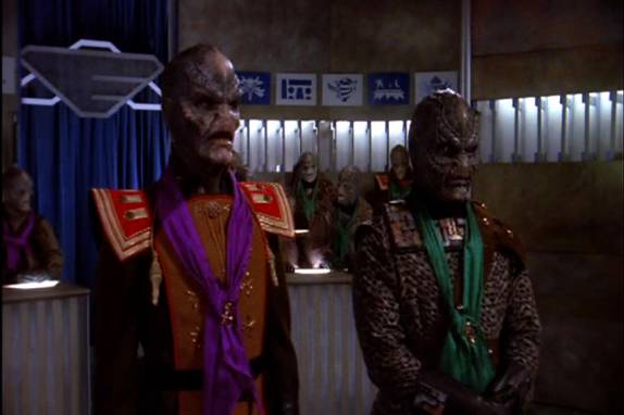 Two Klingon characters from Star Trek: The Next Generation stand in a courtroom-like setting, one wearing a purple scarf and…