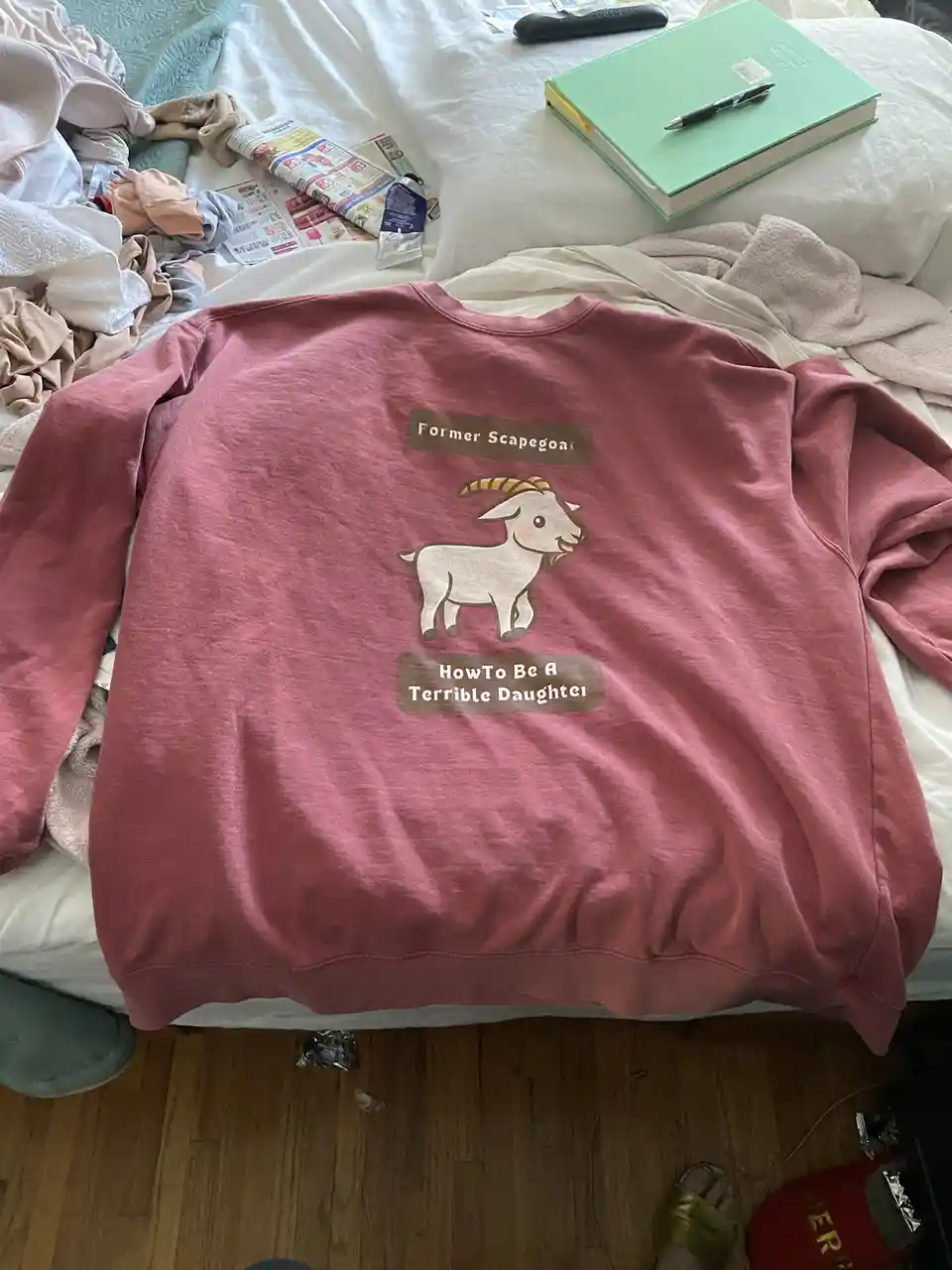A pink sweatshirt laid on a messy bed features a cartoon goat graphic with the text 'Former Scapegoat' above it and 'HowTo…