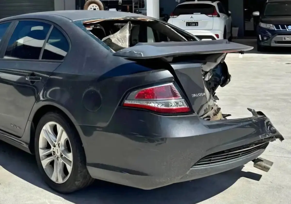 A heavily damaged dark gray Ford Falcon sedan is shown from the rear, with its roof completely torn off and rear bumper…