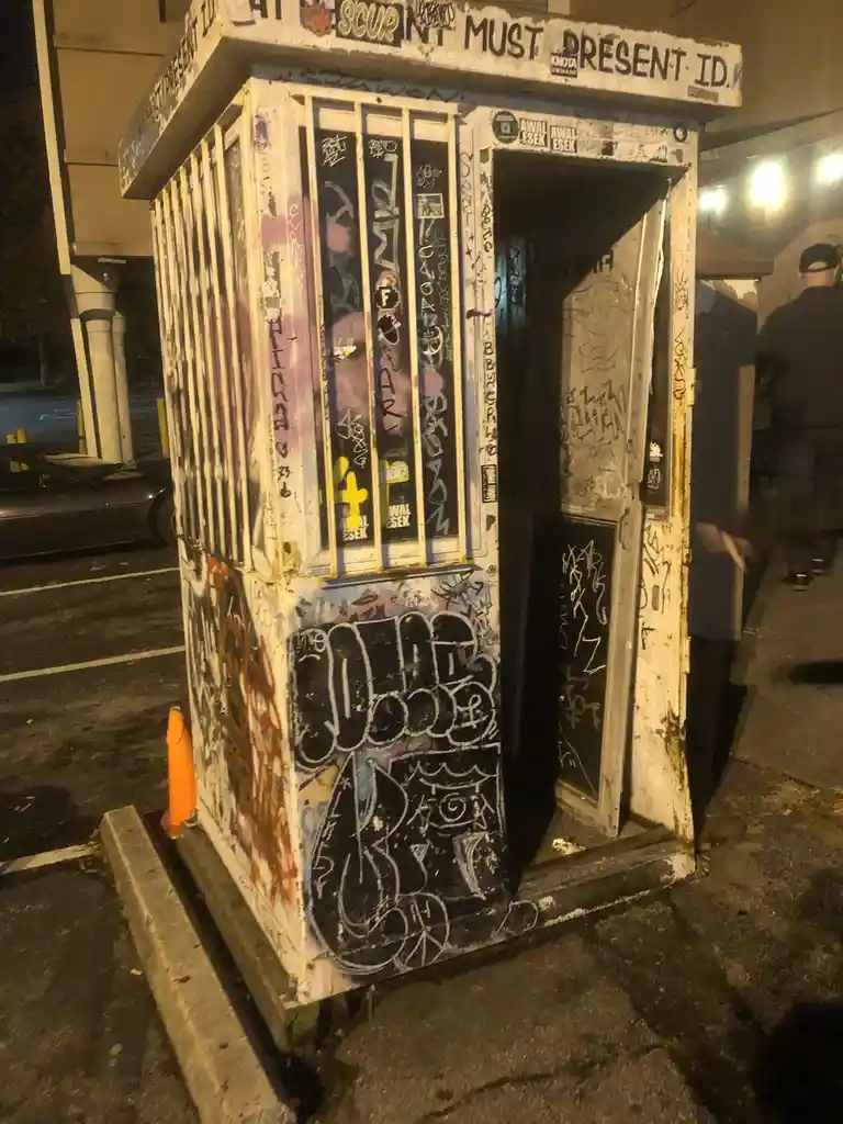 A heavily graffiti-covered, dilapidated white security booth stands in a parking lot at night, its door ajar. The top of the…