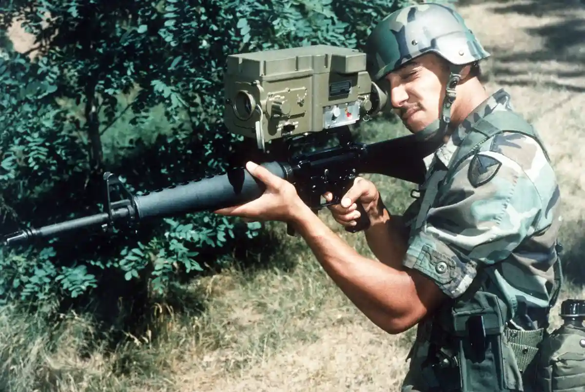 A soldier in camouflage uniform and helmet holds an m16 rifle modified with a large, boxy, olive-drab laser rangefinder or…