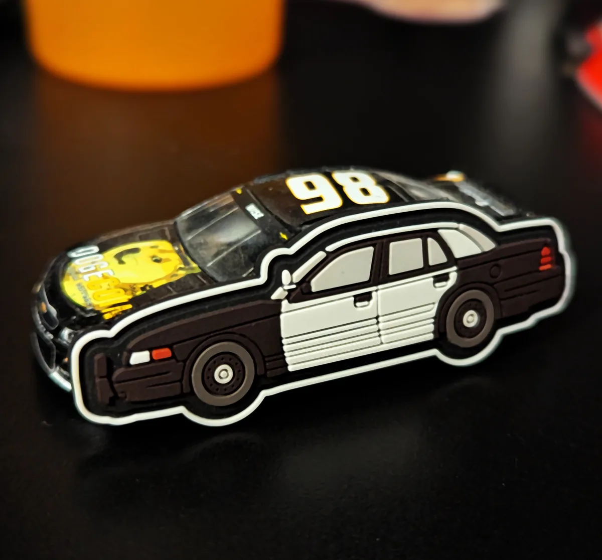 A toy car model resembling a police cruiser with a black-and-white paint scheme, featuring the number 98 on the roof and a…
