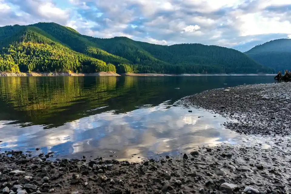 A scenic photograph of a calm lake with forested mountains in the background under a partly cloudy sky. The water is shallow…
