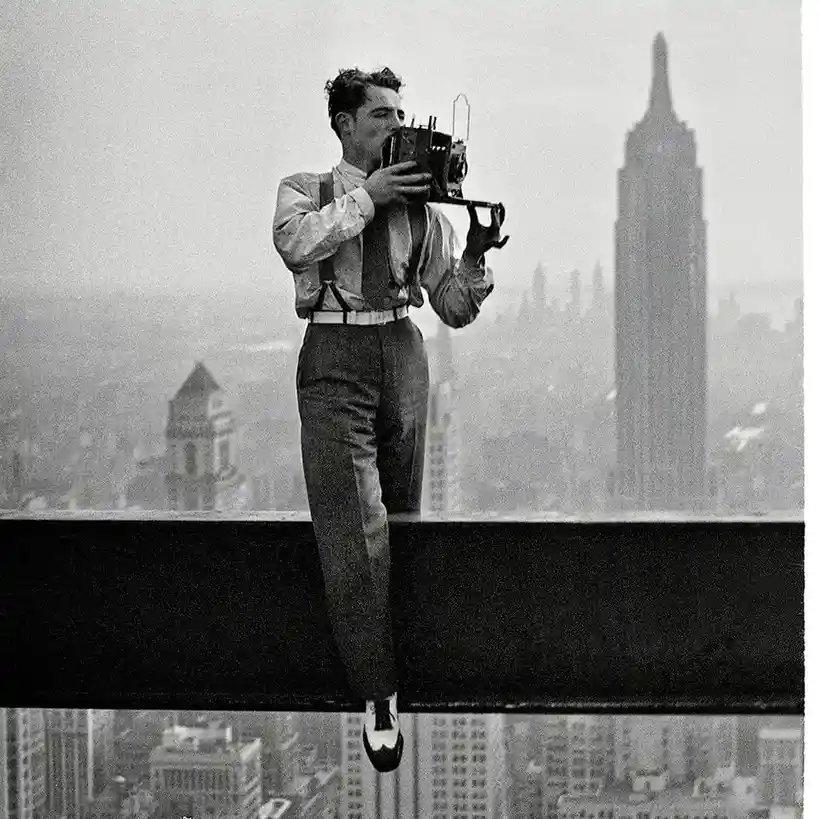 A black-and-white photograph shows a man standing on a narrow steel beam high above a city, holding a large-format camera to…