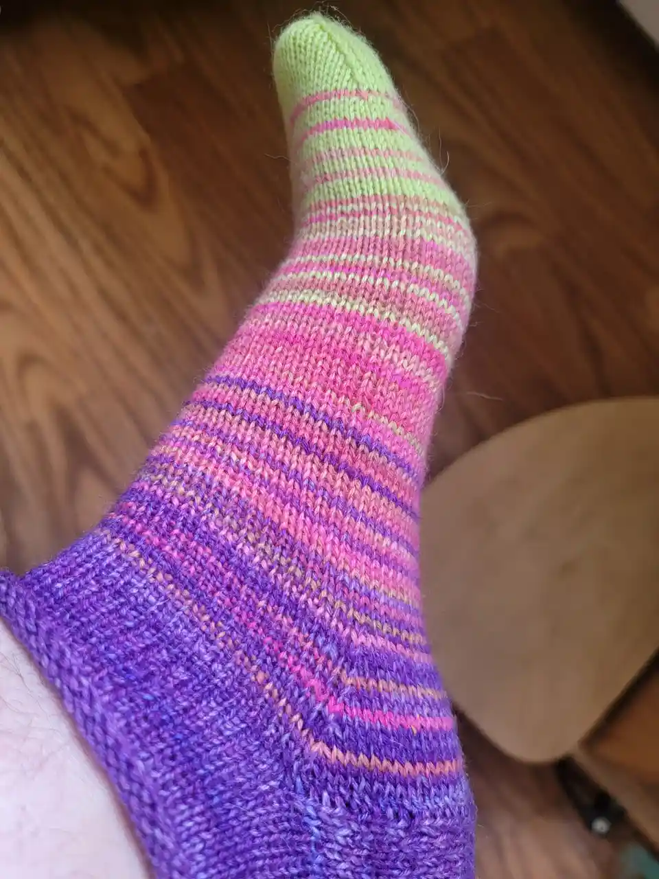 A close-up photo shows a person's foot wearing a hand-knitted sock with a colorful gradient pattern transitioning from lime…