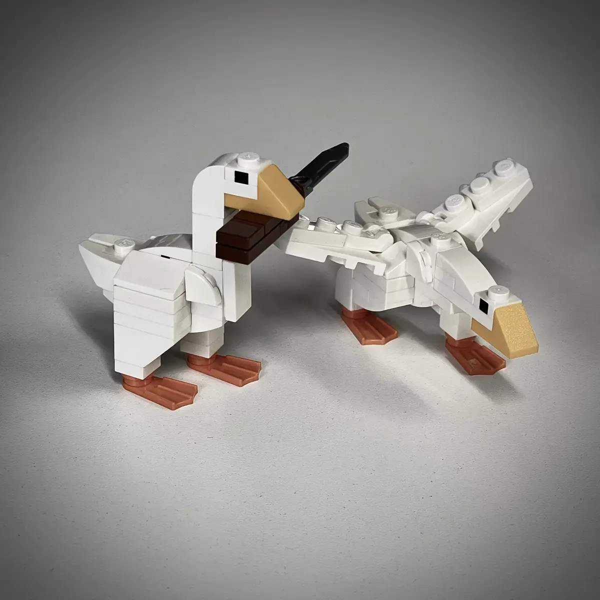 Two white LEGO ducks with tan beaks and orange feet stand side by side on a gray surface. The duck on the left holds a black…