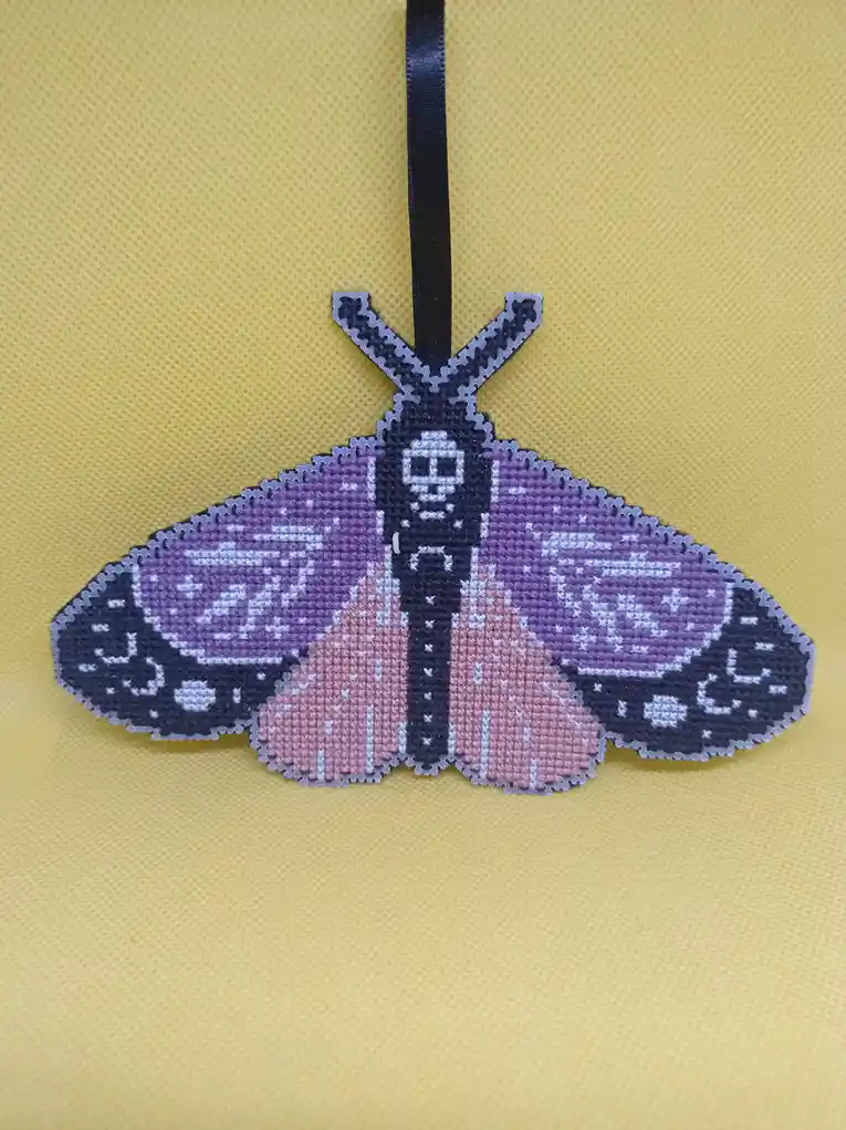 A handcrafted cross-stitch butterfly ornament with purple, pink, and black wings featuring white patterns, hanging by a…