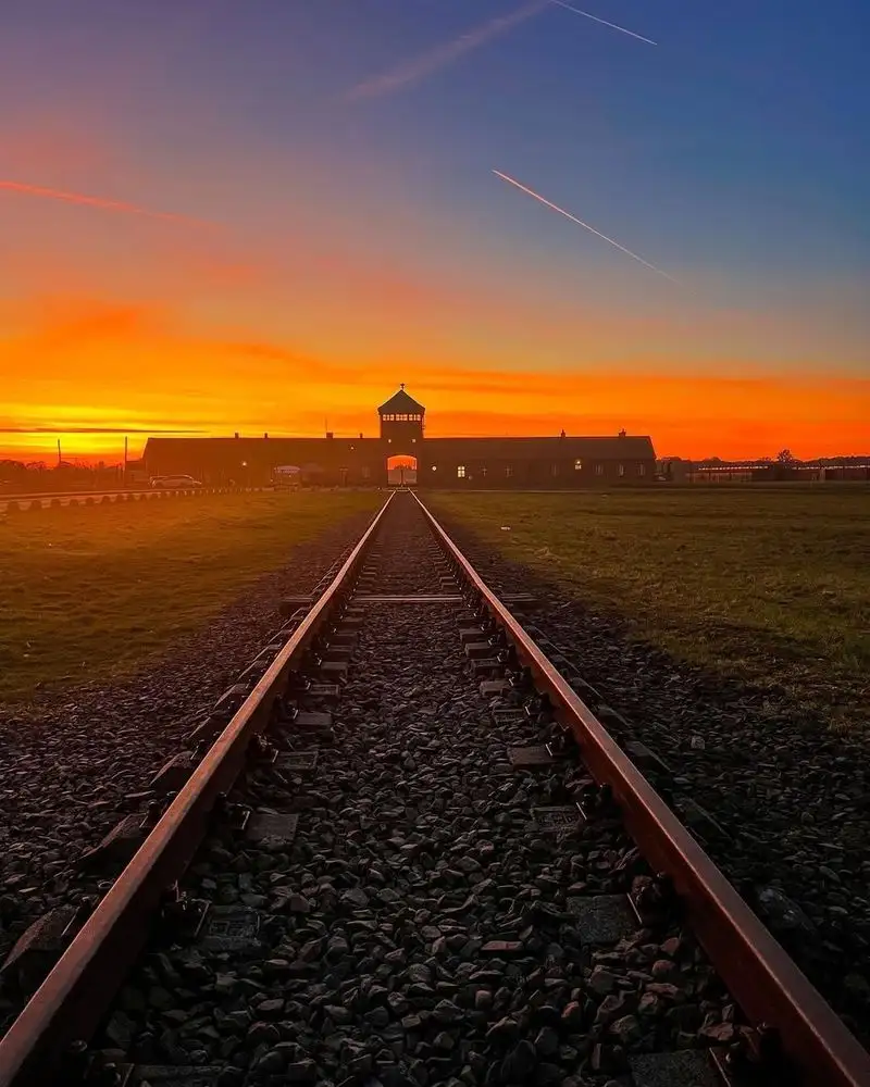 a dramatic sunset photo shows train tracks leading directly toward the iconic gate of the former Auschwitz concentration…