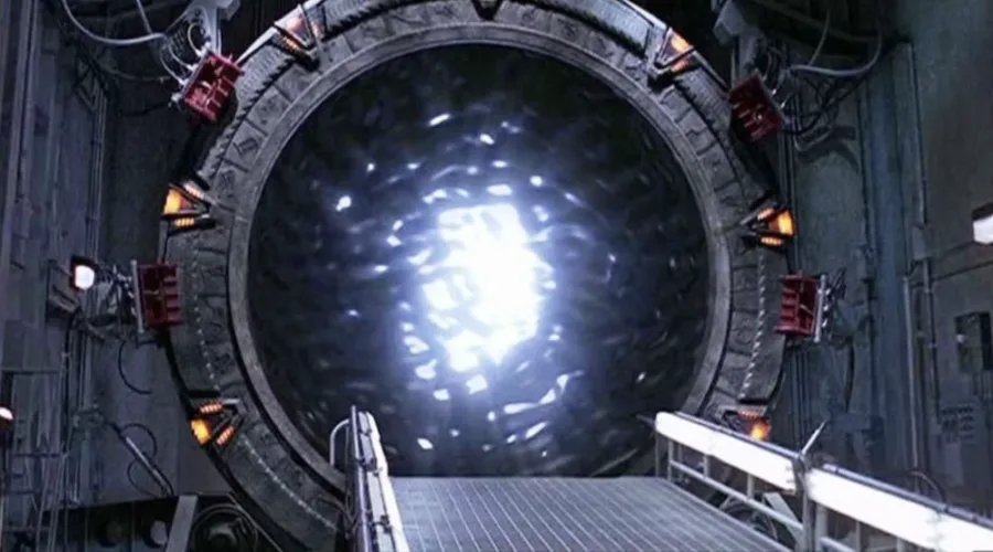 A large, circular, metallic Stargate-like portal emits a bright, swirling light, surrounded by industrial equipment and red…