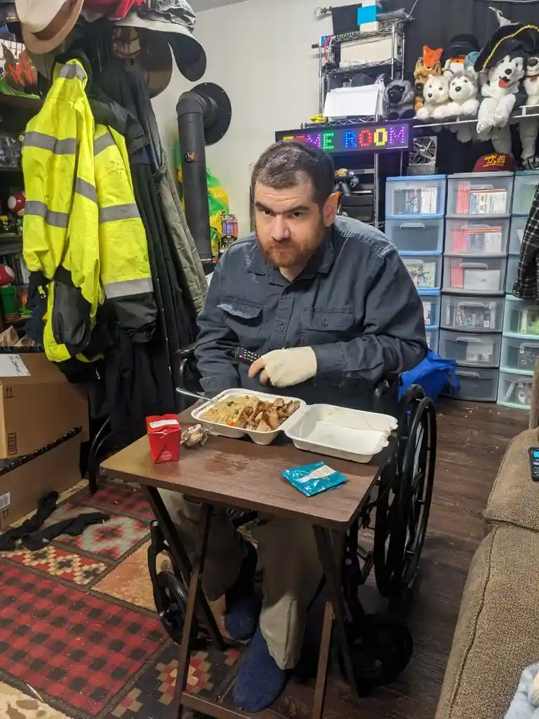 A man with a beard and a cast on his right hand sits in a wheelchair at a small table, holding a remote control. In front of…