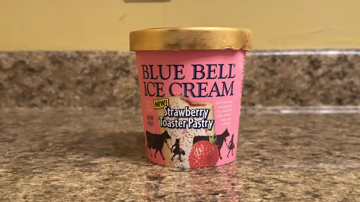 A pint of Blue Bell ice cream labeled 'Strawberry Toaster Pastry' sits on a speckled countertop. The pink container features…