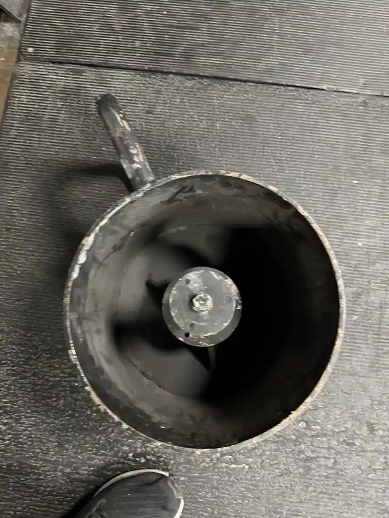 A top-down view of a dirty, black, cylindrical industrial fan or blower with a handle, resting on a textured gray floor. The…
