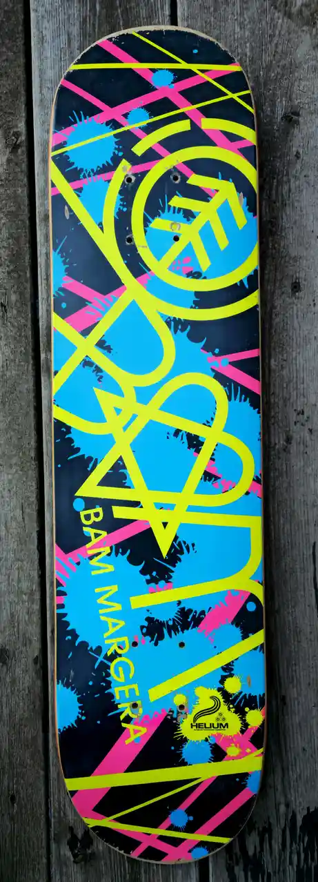 A skateboard deck with a vibrant, abstract design featuring neon yellow, bright pink, and cyan blue splatters and lines on a…