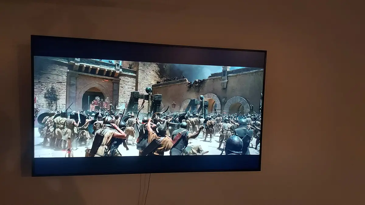 A TV screen mounted on a wall displays a chaotic medieval battle scene with soldiers in armor fighting amidst stone…