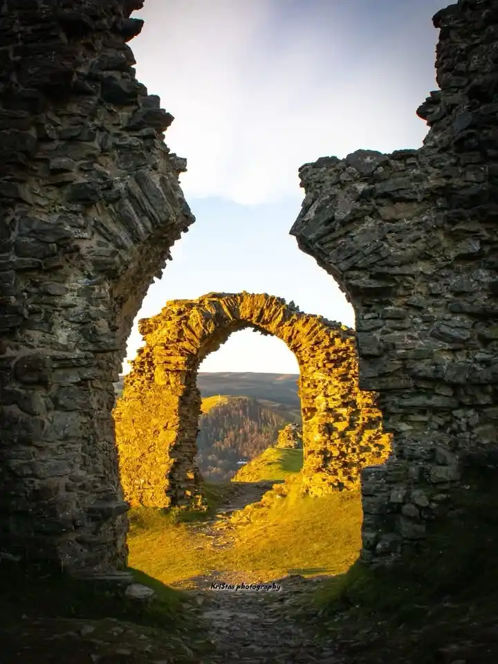 A photograph shows a stone ruin with a broken archway framing another distant stone arch bathed in golden sunlight. The…