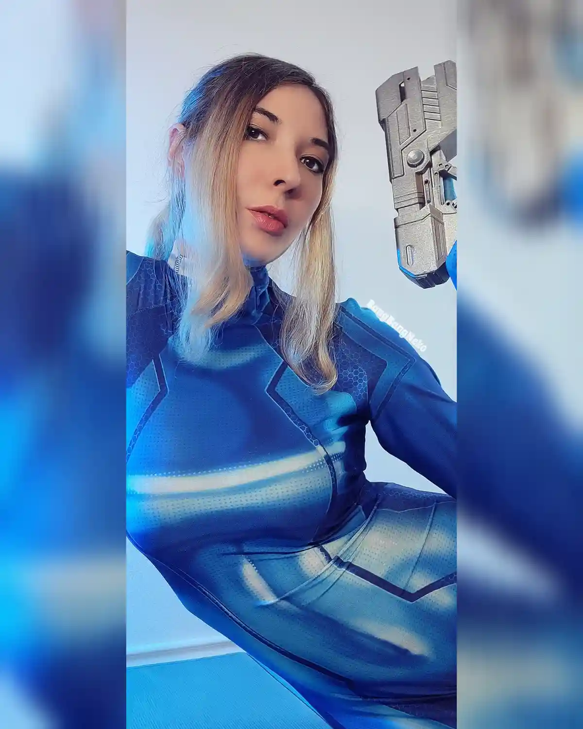 A woman in a form-fitting blue futuristic suit with hexagonal patterns poses while holding a large, angular sci-fi weapon…