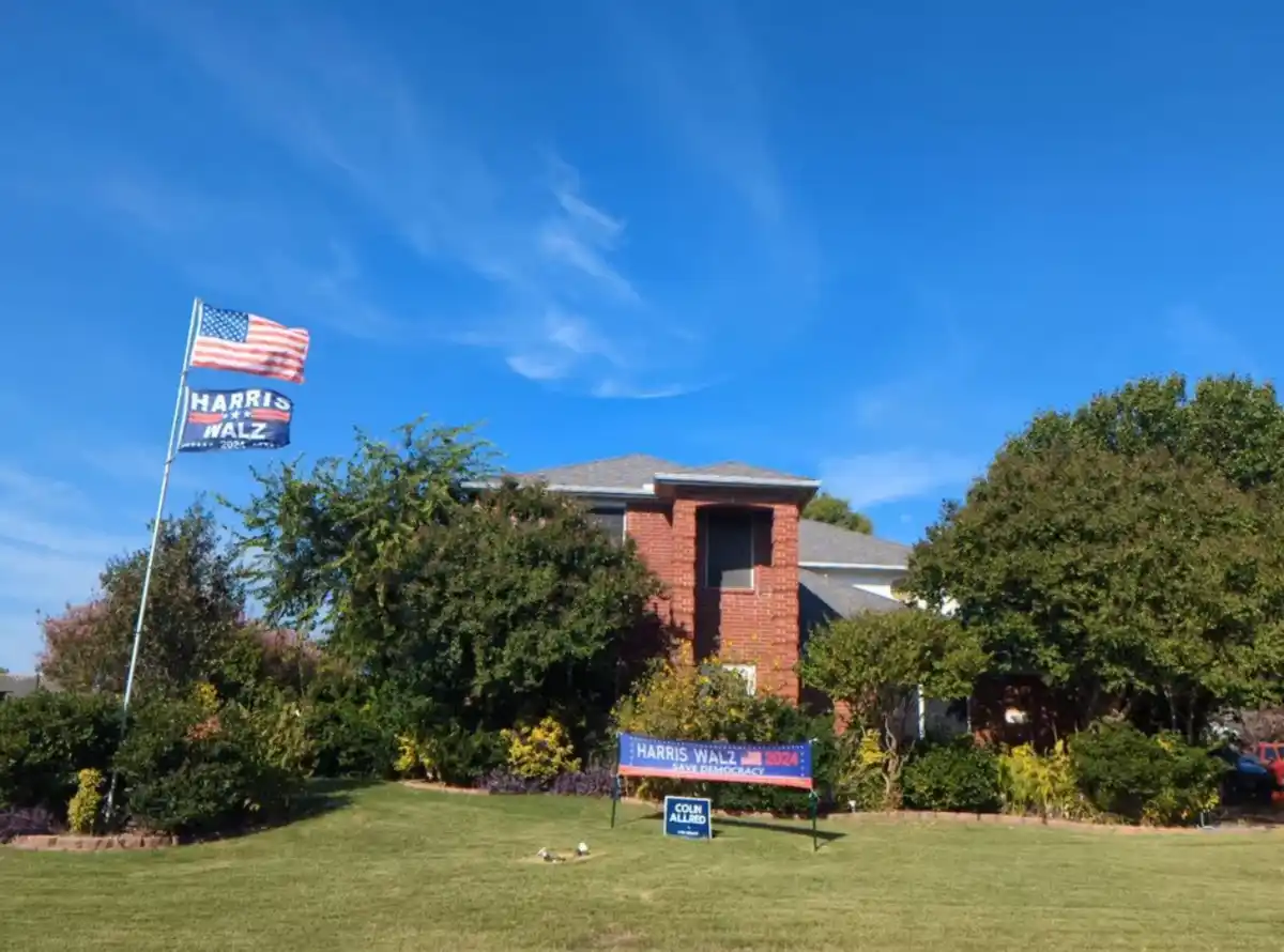 A residential front yard displays multiple 'Harris Walz 2024' campaign signs and banners, including one on a flagpole flying…