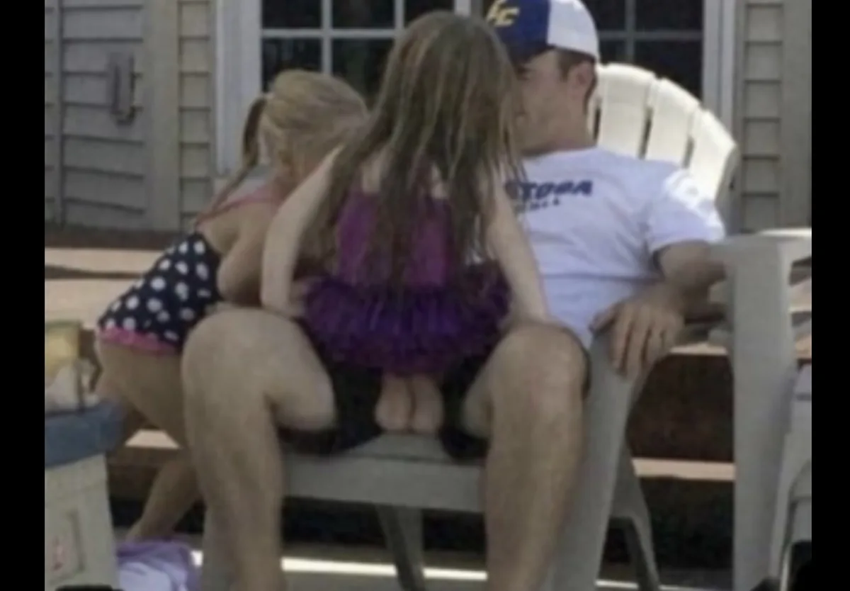 A man in a white t-shirt and baseball cap sits in a white plastic chair outdoors, with two young girls in swimsuits on his…