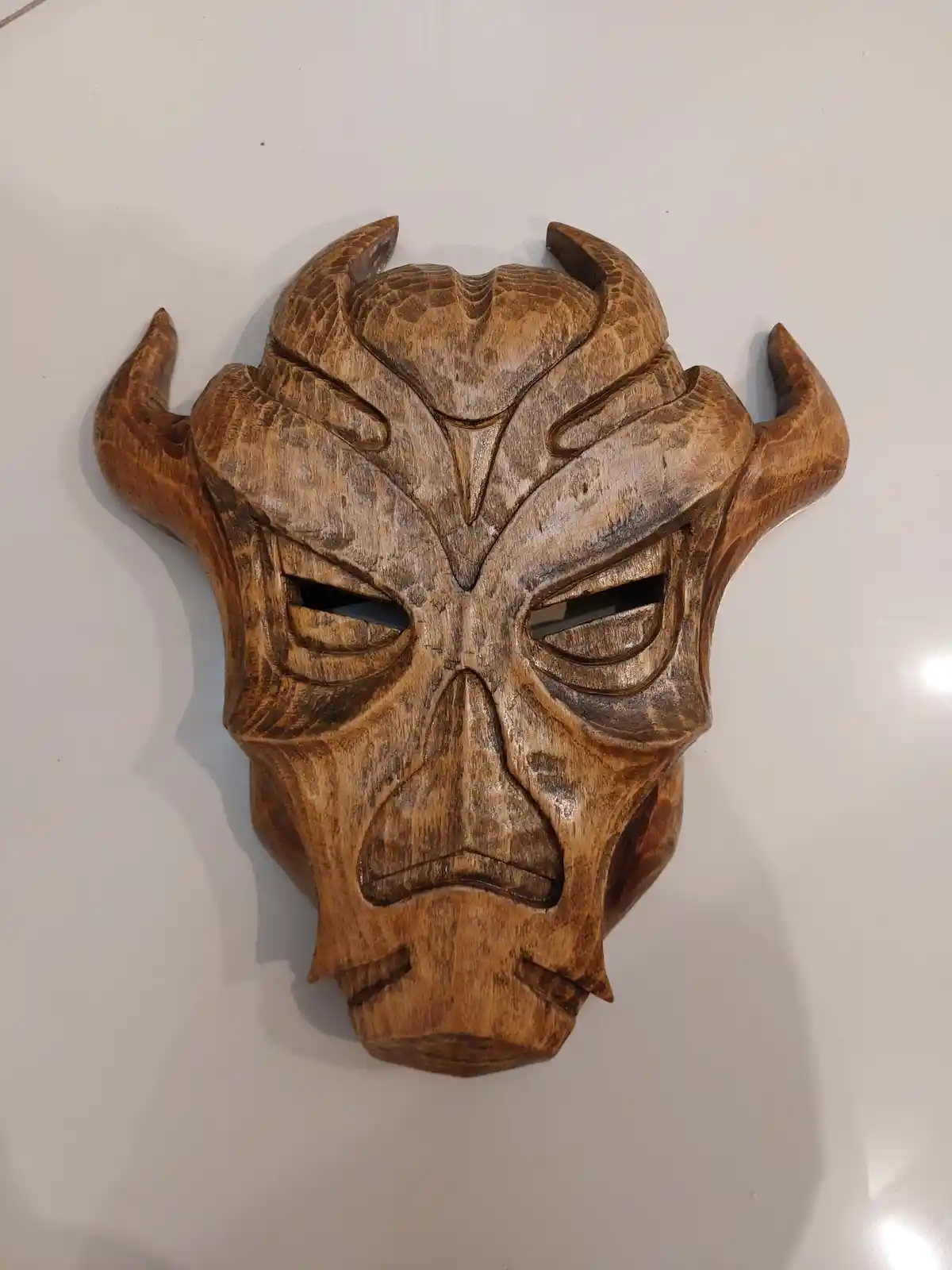 A wooden carving of a demonic mask with horns and an angry expression is mounted on a white wall. The overlaid text reads…