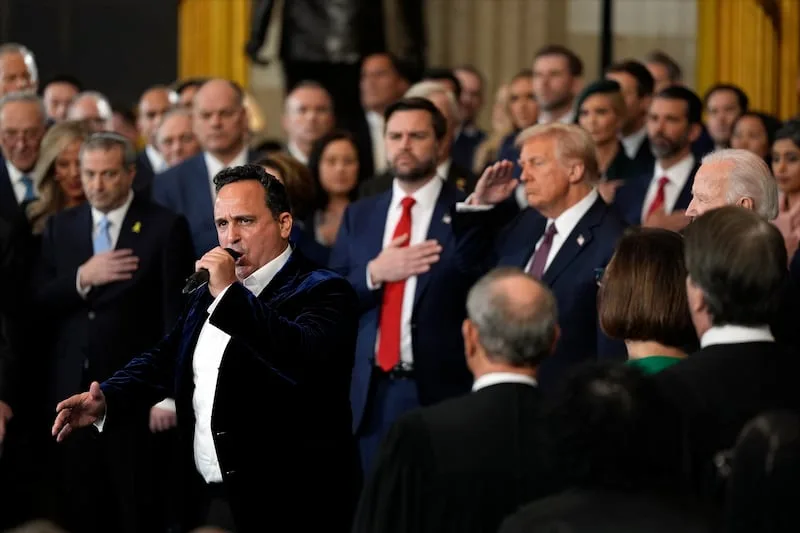 A man in a dark velvet tuxedo jacket and white shirt sings into a microphone while standing in front of a crowd of formally…