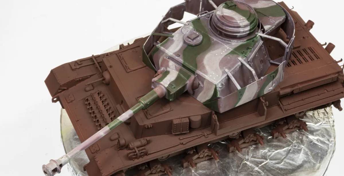 A detailed scale model of a military tank with a camouflage-painted turret and brown hull, resting on a foil-covered base…