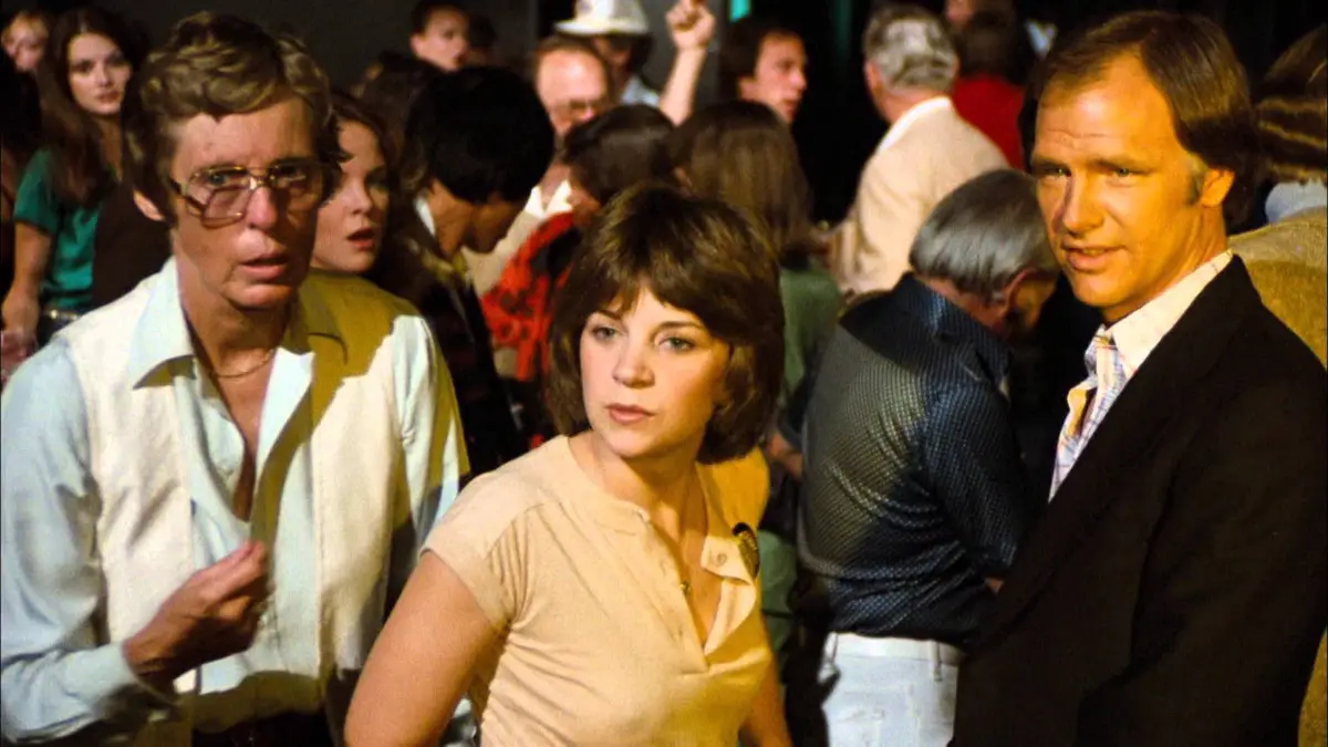 A still from a 1970s film or TV show shows three people in a crowded room. A man on the left with large glasses looks…