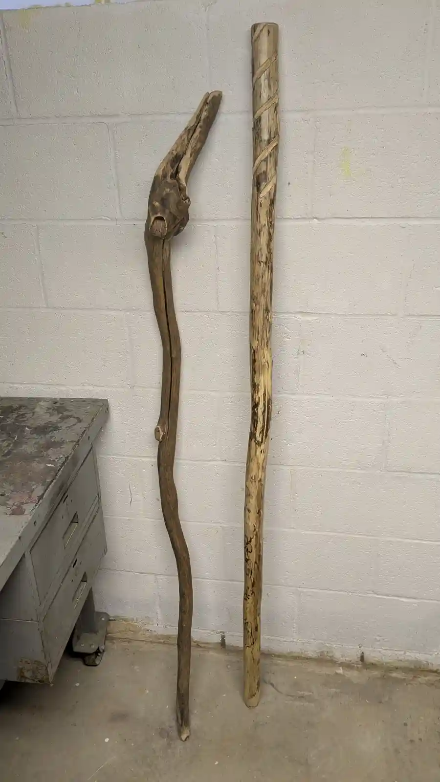 Two wooden walking sticks lean against a white cinder block wall next to a metal filing cabinet. The stick on the left is…