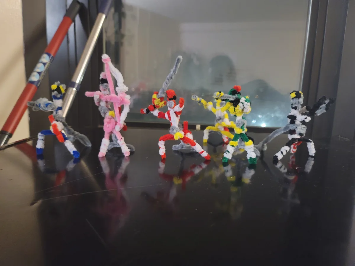 Five handmade figurines crafted from colorful pipe cleaners stand in dynamic battle poses on a reflective black surface…